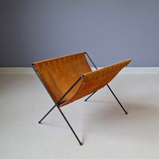 This magazine holder from the 50s/60s features a foldable black metal frame and a rust brown leather (suede) sling. In...
