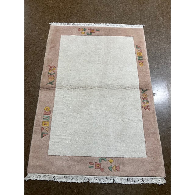 Hand-knotted Nepal wool rug featuring a soft ivory field framed by a wide blush-toned border with abstract geometric...
