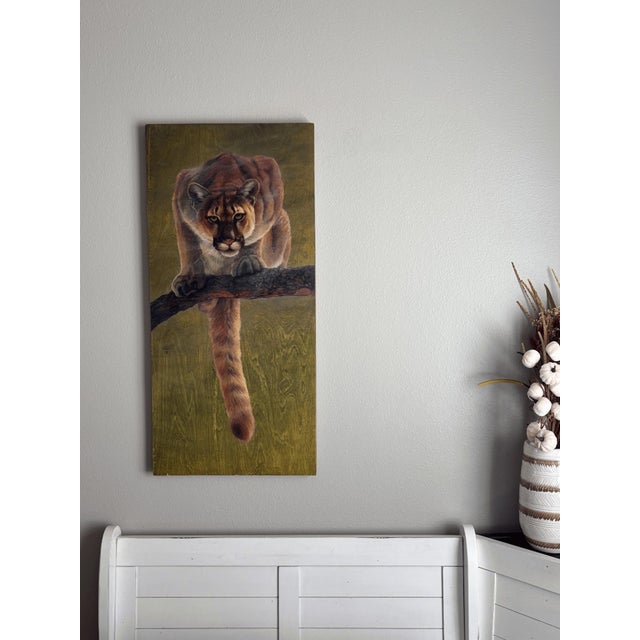 This oil painting depicts a poised mountain lion, its intense gaze focused intently as it crouches on a branch, ready to...