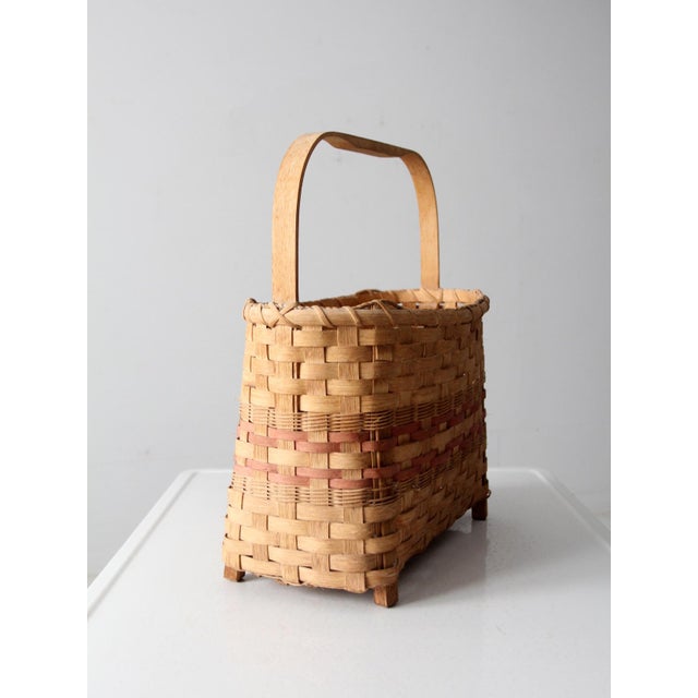 A vintage American splint basket featuring a footed base, arched handle, and classic flat-splint weave accented with soft...