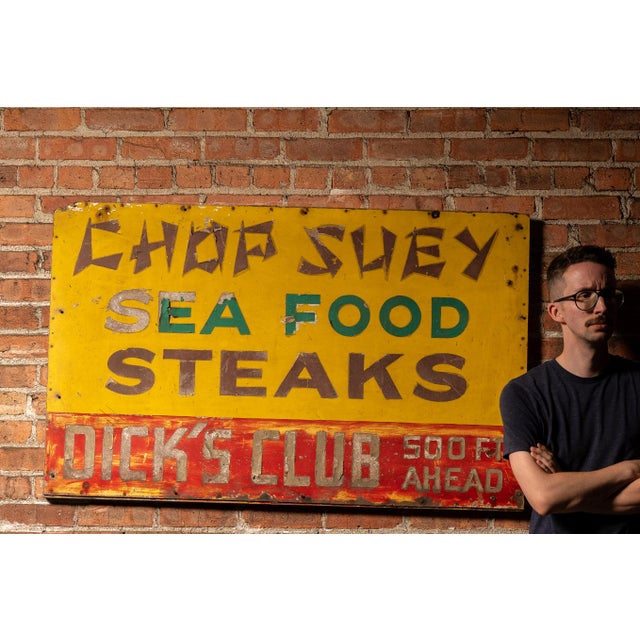 Painted roadside metal sign promoting Chop Suey, Sea Food, and Steaks, with a bold red banner along the bottom reading...
