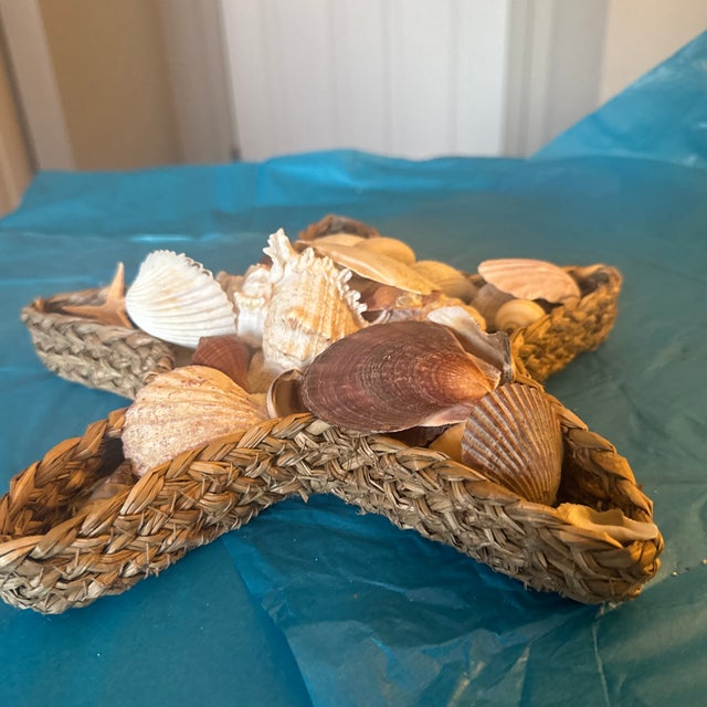 This decorative starfish-shaped woven basket is filled to the brim with a diverse collection of natural seashells, resting...