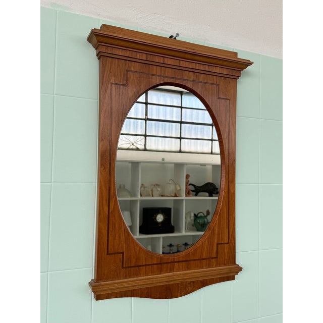 An elegant oval mirror with a wooden frame featuring a contoured top panel reminiscent of vintage mirrors. Made by an...