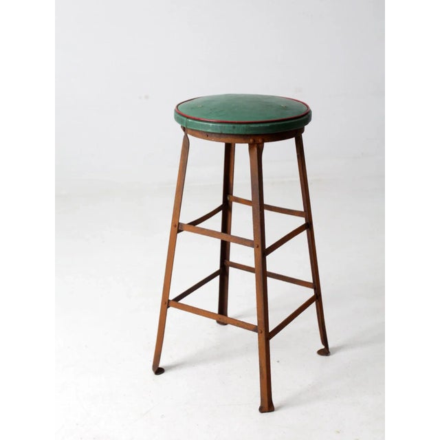 This is a vintage industrial work stool. The tall metal frame stool features a round green upholstered seat with red trim.