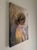 This oil on linen painting captures a contemplative figure draped in a soft yellow garment, her gaze directed downward and...
