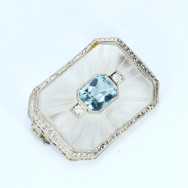 This brooch features a central aquamarine stone, cut in a mixed style, displaying a captivating blue hue. Flanking the...