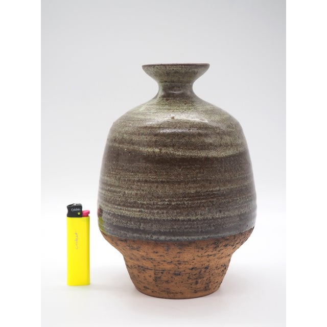 A hand-thrown ceramic vase with a gently rounded body, narrow neck, and shallow mouth. The glaze's earthy tones of green...