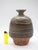 A hand-thrown ceramic vase with a gently rounded body, narrow neck, and shallow mouth. The glaze's earthy tones of green...