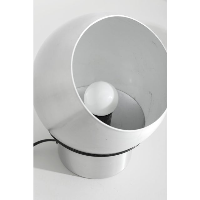 A sculptural aluminum table lamp defined by its spherical shade and open aperture that frames the exposed bulb within. The...