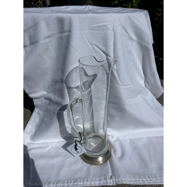 Vintage tall clear glass pitcher featuring an elegant elongated form, applied curved handle, and decorative silver plate...