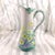 This is a pitcher that is pink at the top, purple at the bottom and has a mint green shaped rim and handle. The handle is...
