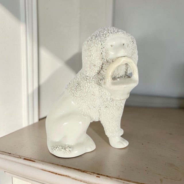A miniature Victorian Staffordshire poodle modelled seated and holding a loop-handled basket in its mouth. The compact...