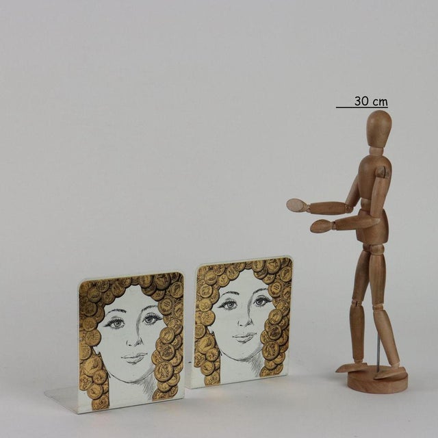 Pair of bookends with silkscreen depicting a face with coins. Manufacture mark under the base. This piece is attributed to...
