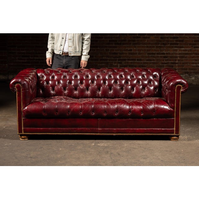 Classic tufted Chesterfield sofa upholstered in oxblood red leather with deep buttoned cushioning, rolled arms, and brass...