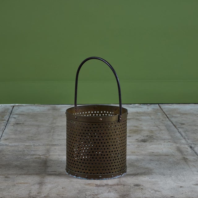 Brass pail with curved handle. This piece features a round perforated body with rivets at both the top and bottom of the...