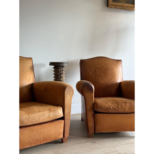 A pair of French leather chairs in a warm honey tan, with arched moustache backs, rolled arms, and loose seat cushions....
