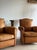 A pair of French leather chairs in a warm honey tan, with arched moustache backs, rolled arms, and loose seat cushions....