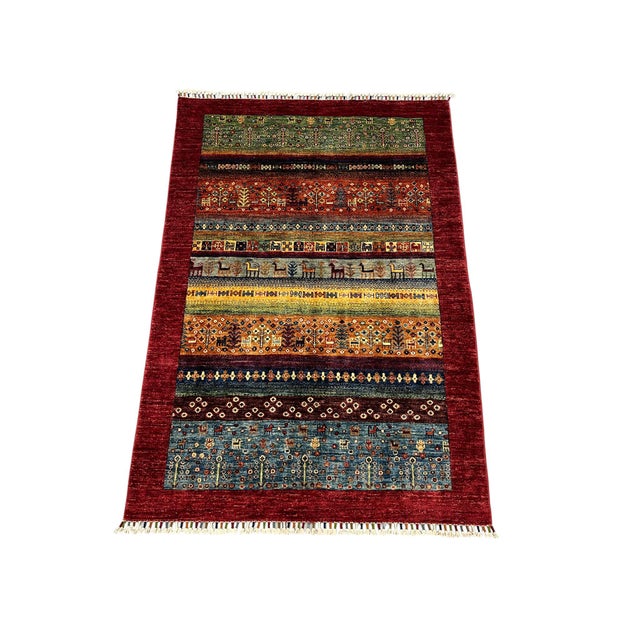 This khorjin saddlebag-inspired area rug features rows of small animal figures, trees, and patterned bands. A broad deep...