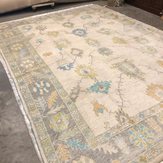 A Turkish Oushak rug featuring an ivory field a with large-scale floral motifs in soft turquoise, muted orange, and gentle...