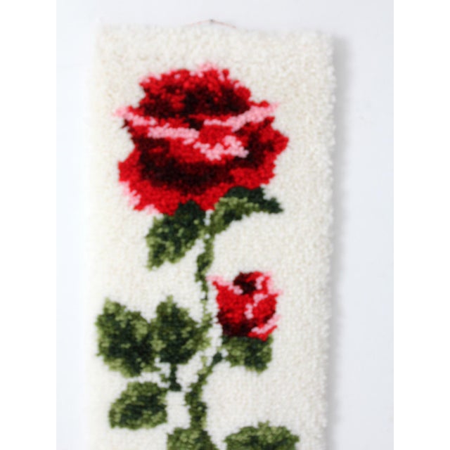 A vintage Shillcraft latch hook wall hanging featuring a bold red rose set against a soft ivory ground. The raised pile...