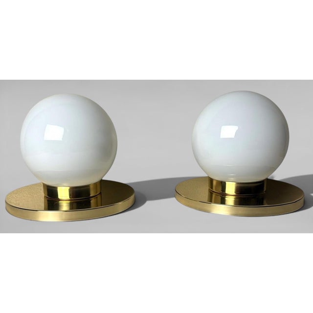 A refined pair of table lamps featuring opal glass shades and sturdy circular brass bases, through which the lamp holder’s...