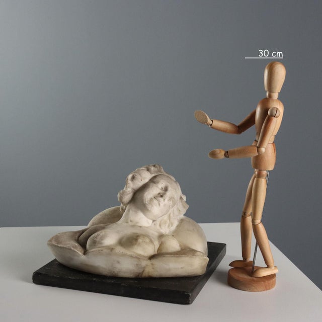 The marble bust depicts a young woman with a smiling face. Her curly hair wraps around her face, and her long hair is...