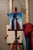 This oil painting features a profile of a man draped in a vibrant blanket, characterized by bold red, white, and blue...
