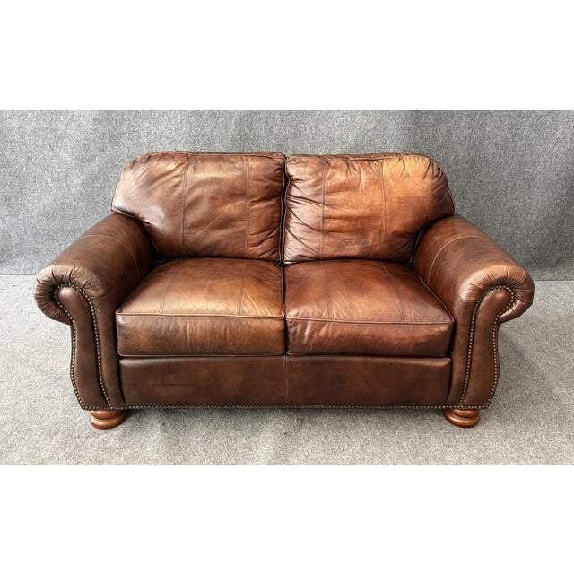 This Thomasville brown leather loveseat features a classic two-seat design with plush cushions, rolled arms, and nailhead...