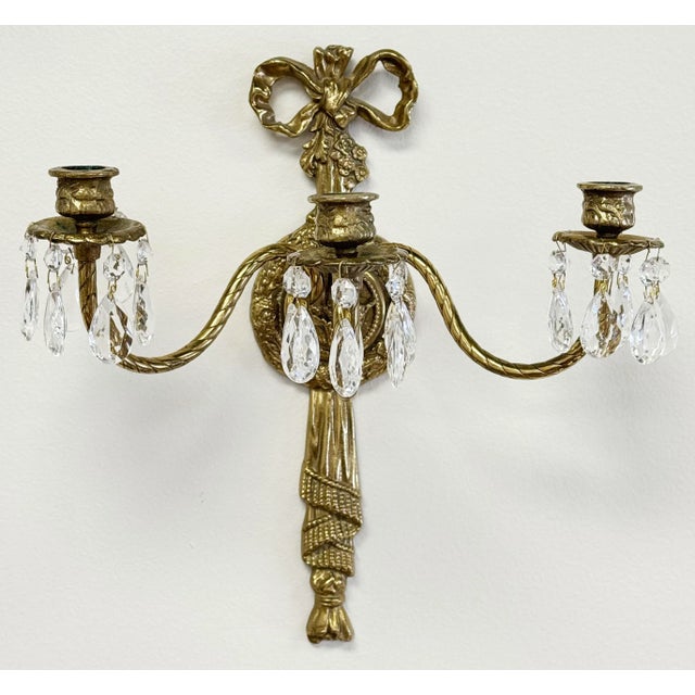 Pair of vintage French Empire brass three arm wall candle sconces with crystal drops. Featuring stunning timeless ribbon...