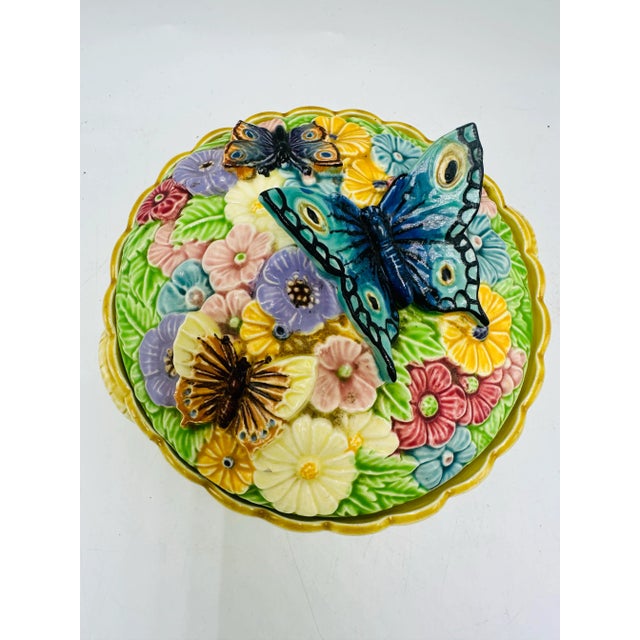 This is a vibrant ceramic dish with a lid, featuring detailed floral and butterfly motifs. The lid is adorned with...