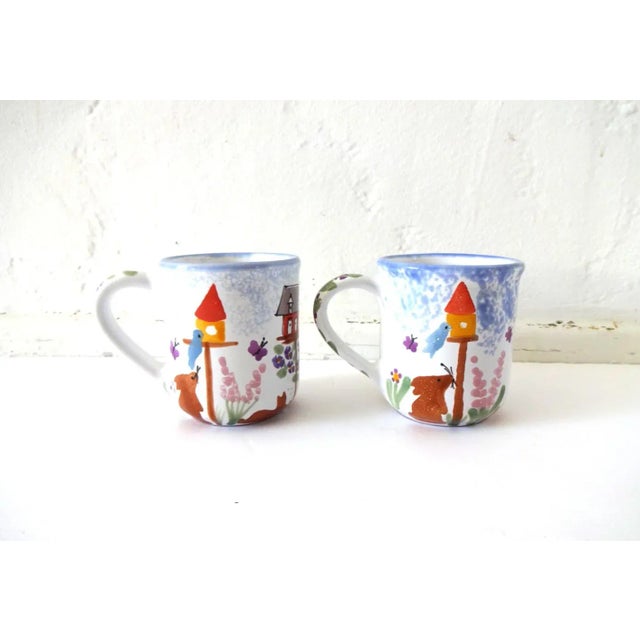 A pair of vintage mugs, hand-painted with bunnies, birds, bird houses, flowers, and a speckled blue sky. Purple flowers...