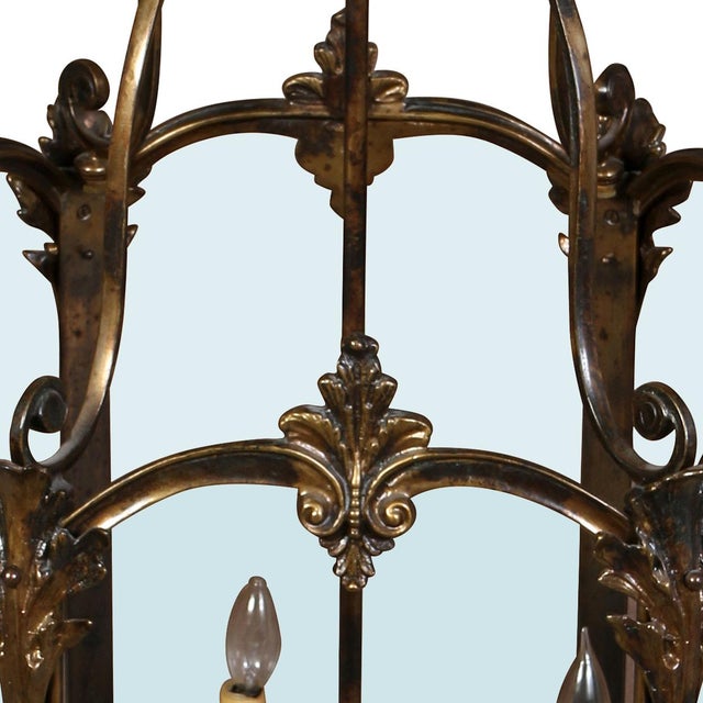 A lantern chandelier crafted in brass, featuring nine candle style lights enclosed within tall, gracefully curved glass...