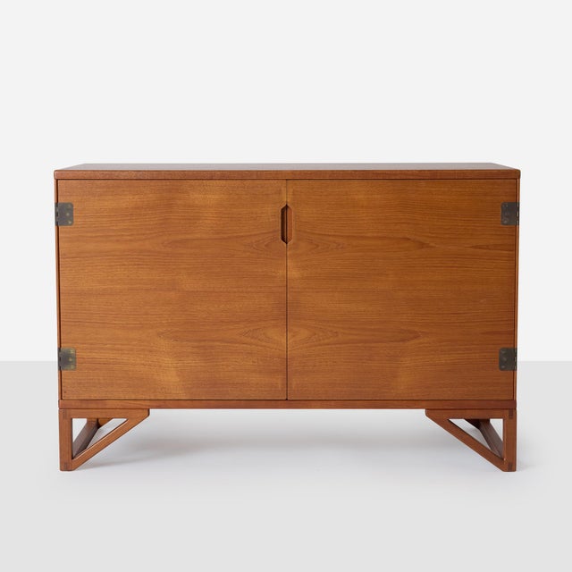 This teak sideboard sits on an angular base and features two internal shelves, three drawers and patinated brass hinge...