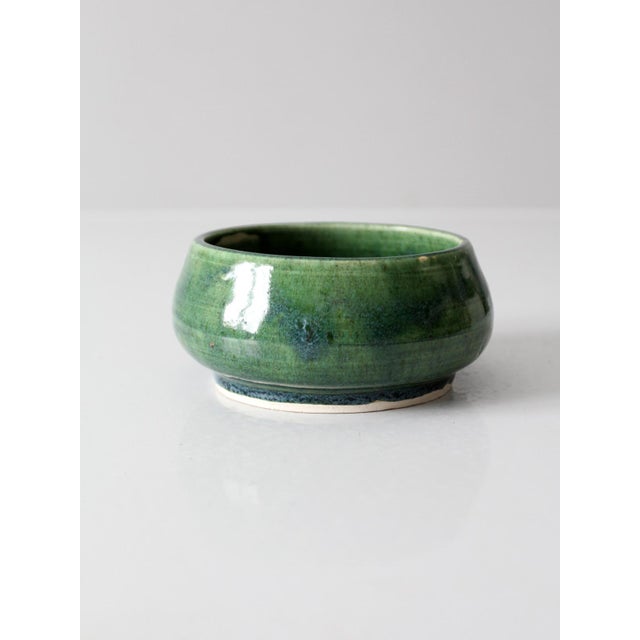 This vintage ceramic bowl features a layered green glaze that shifts in tone across its rounded form. A contrasting...