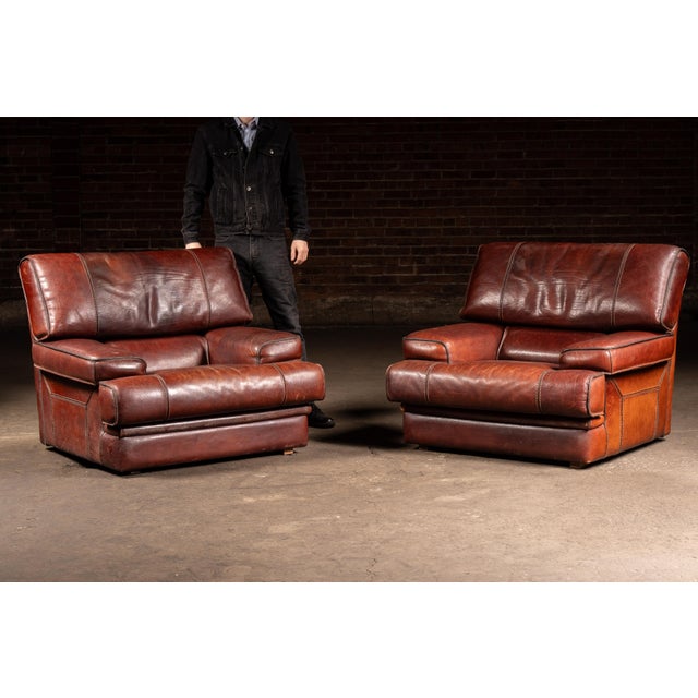 Pair of oversized leather lounge chairs with broad padded arms, deep seat cushions, and low block wood feet. Subtle panel...