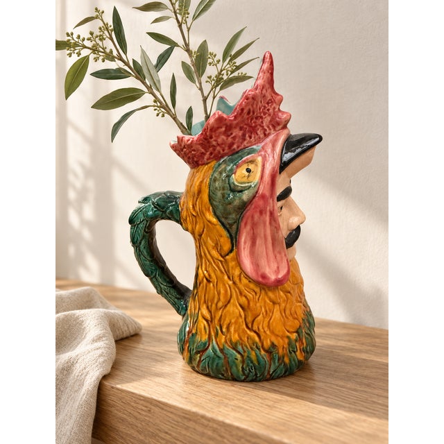 A polychrome glazed ceramic pitcher in the shape of a rooster's head, featuring bold colors: a nuanced red comb, finely...