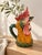 A polychrome glazed ceramic pitcher in the shape of a rooster's head, featuring bold colors: a nuanced red comb, finely...
