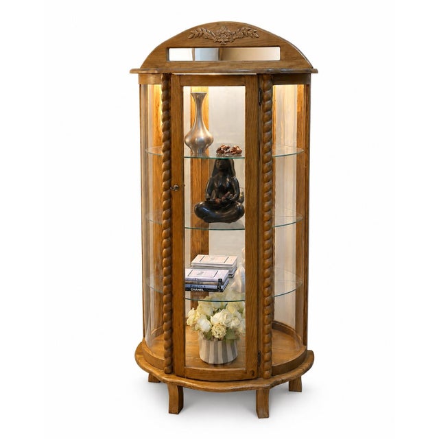 A composed vintage curio cabinet in warm oak, defined by its curved glass silhouette, twisted columns, floral carving,...