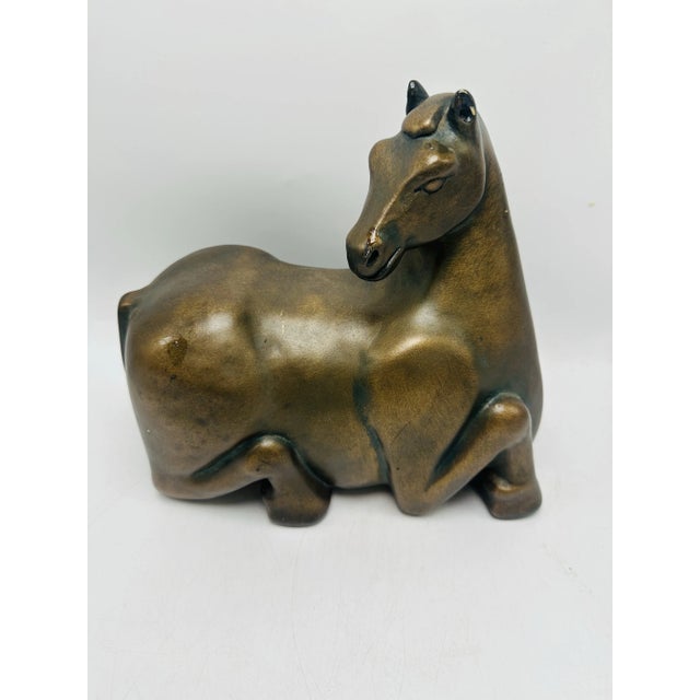 This piece is a sculpture of a reclining horse, crafted with a smooth, bronze-like finish. The stylized design highlights...