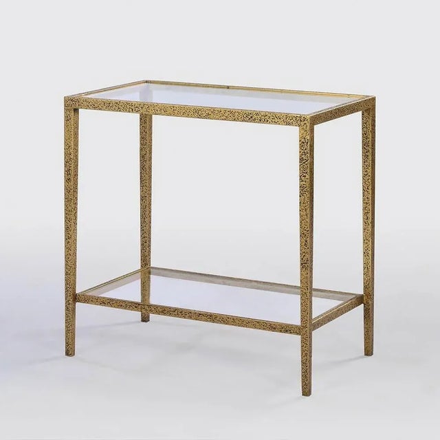 A modern hammered narrow rectangle side table, with clear tempered glass top and shelf, has a “gold leaf” finish on cast...