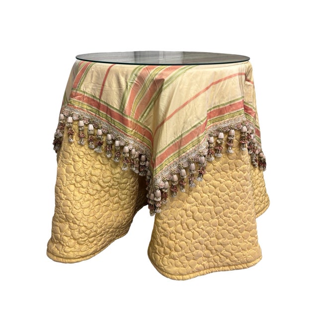 Round side table with a hidden wooden base layered with a floor-length quilted yellow skirt, a striped overlay with...