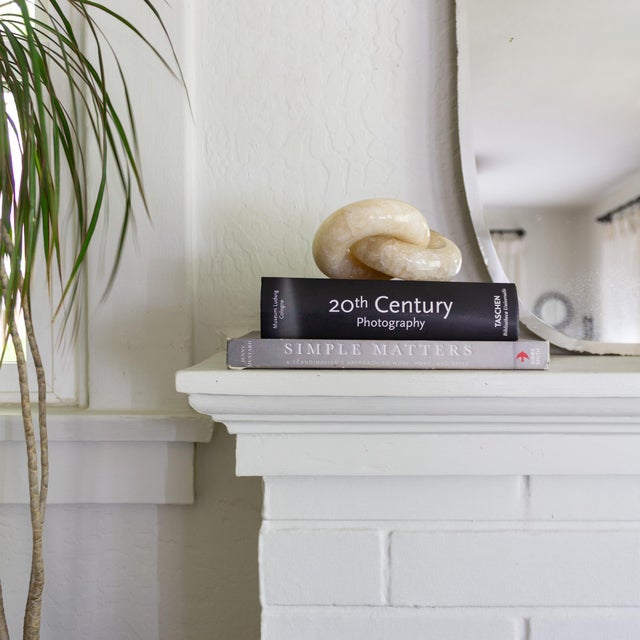 An onyx marble knot sculpture composed of two interlocking rings in soft tones of cream, peach, and taupe. The sculpture...