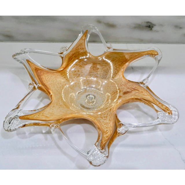 A striking amber and clear art glass centerpiece bowl featuring a dramatic six-point starburst silhouette. Warm honey-...