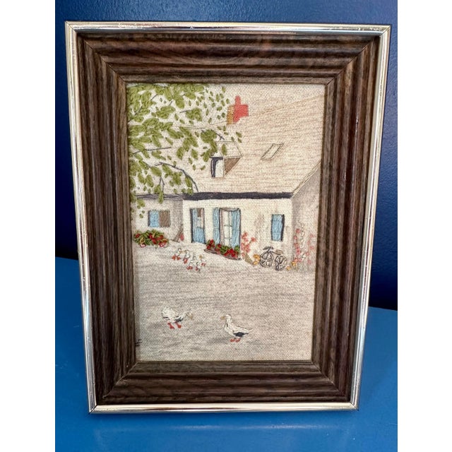 Hand embroidered cottage scene depicting a countryside cottage with geese, a bicycle, flowers, and a cascading tree....