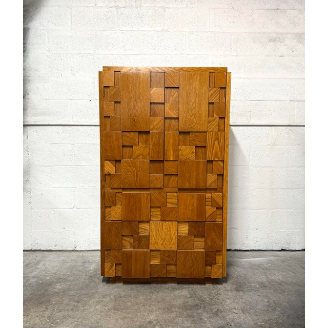 Vintage Lane armoire featuring a sculptural patchwork facade of raised geometric wood panels. A warm honey finish...