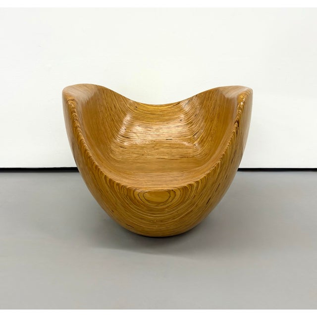 A sculptural carved wood chair featuring a deeply contoured seat, sweeping raised sides, and a rounded base. Warm honey...