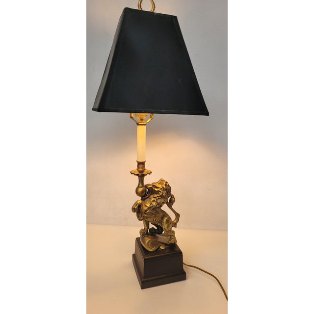 Late-20th-century brass-tone lion table lamp with a black hardback shade, reflective gold lining, and twin-leaf finial. It...