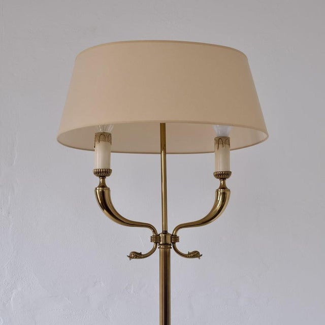 Neoclassical brass floor lamp attributed to maison jansen, featuring twin curved candle arms with dolphin-head details....