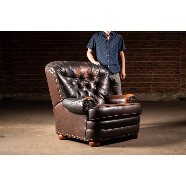 Classic English club chair upholstered in dark brown leather with a deeply button-tufted back, rolled arms, and oversized...