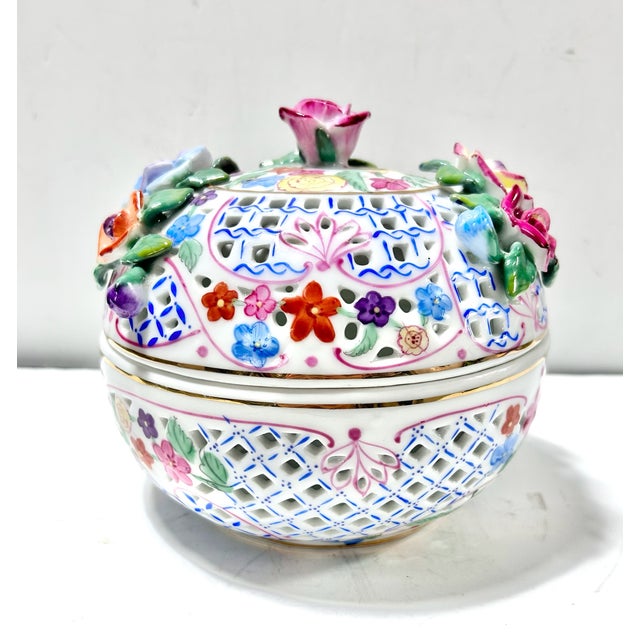 A decorative pierced porcelain lidded jar adorned with vibrant hand-applied roses and colorful floral motifs. The domed...